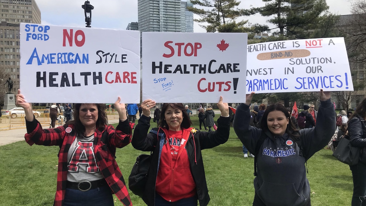Trois manifestantes brandissent chacune une affiche invitant le premier ministre Doug Ford à cesser les coupes en santé.