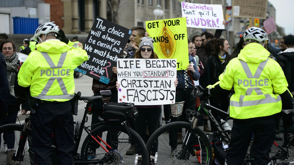 Des policiers bloquent, avec leur bicyclette, le passage à des manifestants tenants des pancartes sur lesquelles sont écrits des slogans pro-choix.