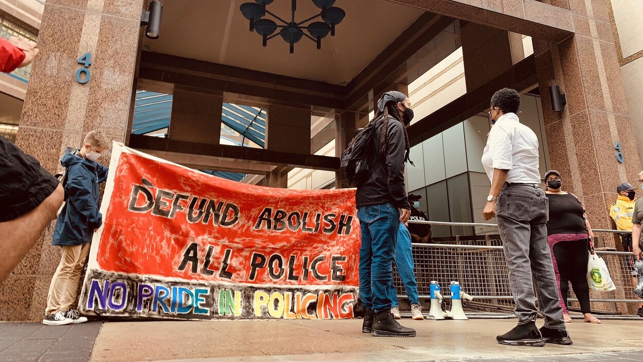 Des manifestants se trouvent devant le Commissariat du Service de police de Toronto et brandissent une affiche sur laquelle il est inscrit « Defund abolish all police ».