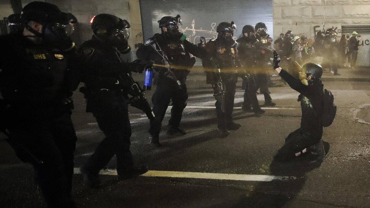 Un manifestant à genoux devant des policiers en rang, dont un l'asperge de poivre.