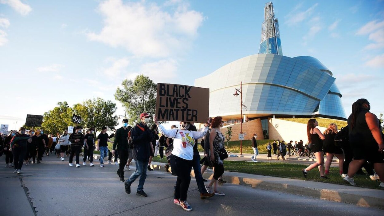 Des personnes marchent dans une rue en tenant des affiches, devant le Musée canadien pour les droits de la personne.