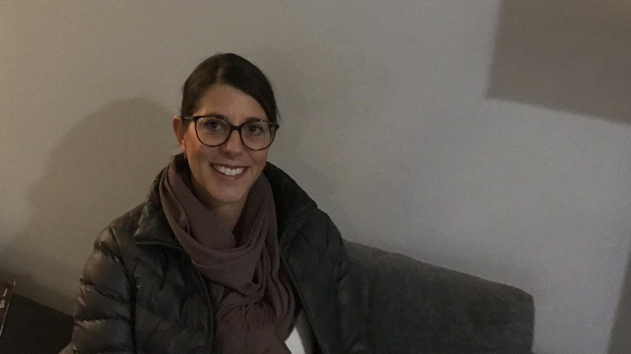 Une femme est assise sur un fauteuil gris devant un mur où est inscrit CBC, Radio-Canada.