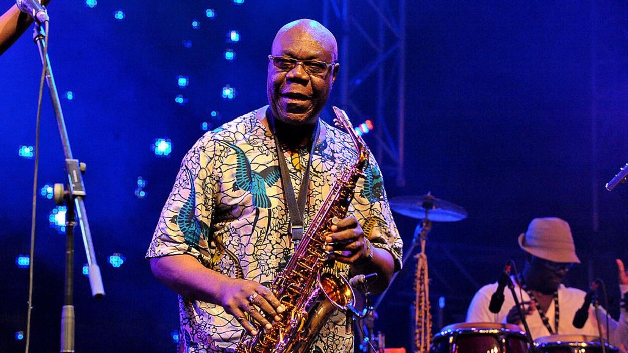 Manu Dibango est sur scène et tient un saxophone dans ses mains.