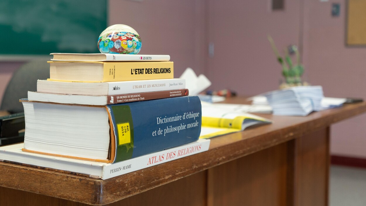 Une pile de manuels scolaires, un dictionnaire et un atlas sur un bureau en bois dans une salle de classe.
