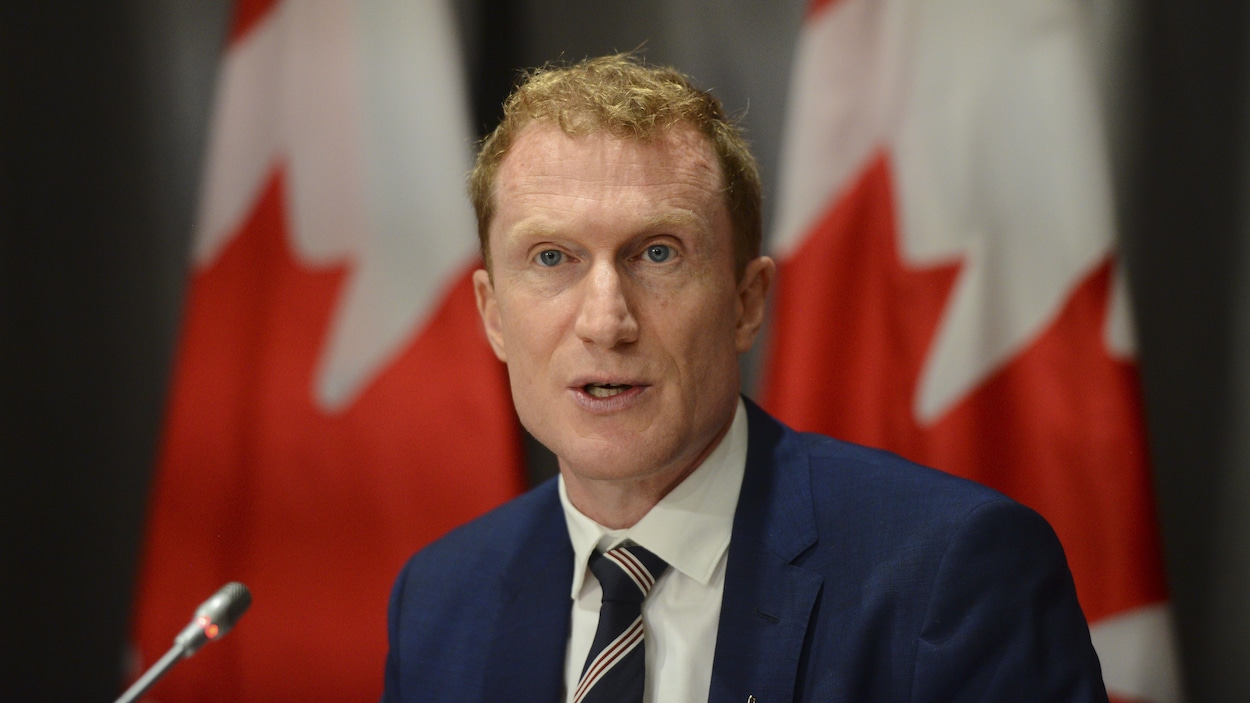 Marc Miller en conférence de presse devant deux drapeaux du Canada.