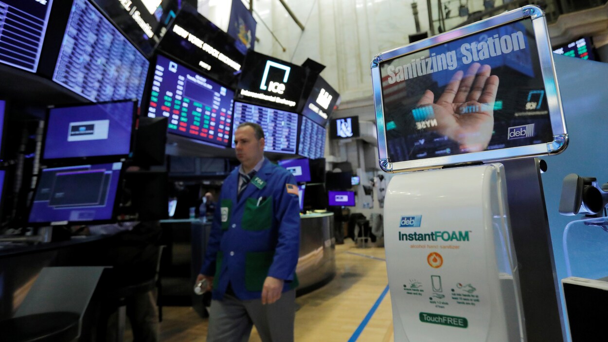 Un distributeur de désinfectant pour les mains est installé à la Bourse de New York; à proximité, un homme marche sous les écrans.