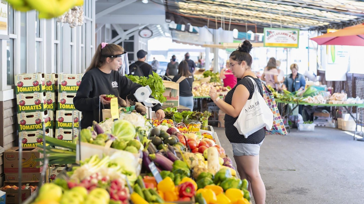 Dans un marché public, une femme fait ses achats devant un étal de légumes, servie par une employée.