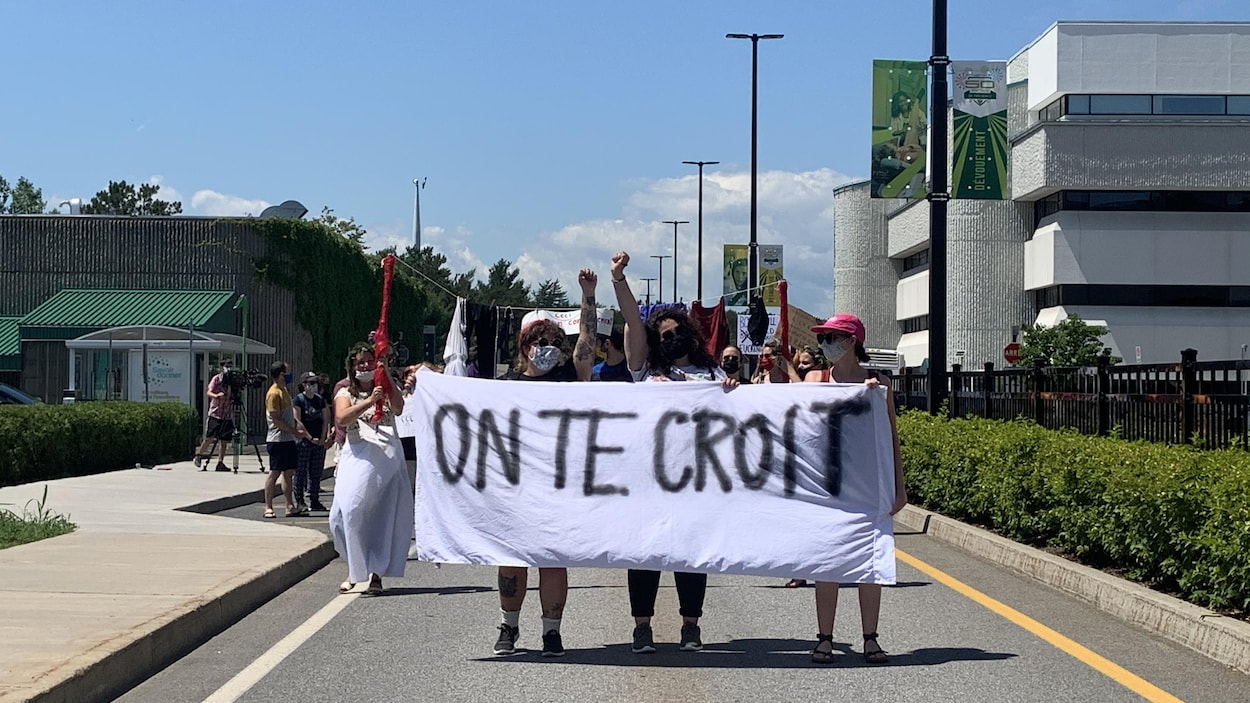 Des manifestantes masquées, poing levé, qui tiennent une banderole sur laquelle est écrit : "On te croit". Elles marchent tout près du lieu de départ, sur le campus de l'UQTR.