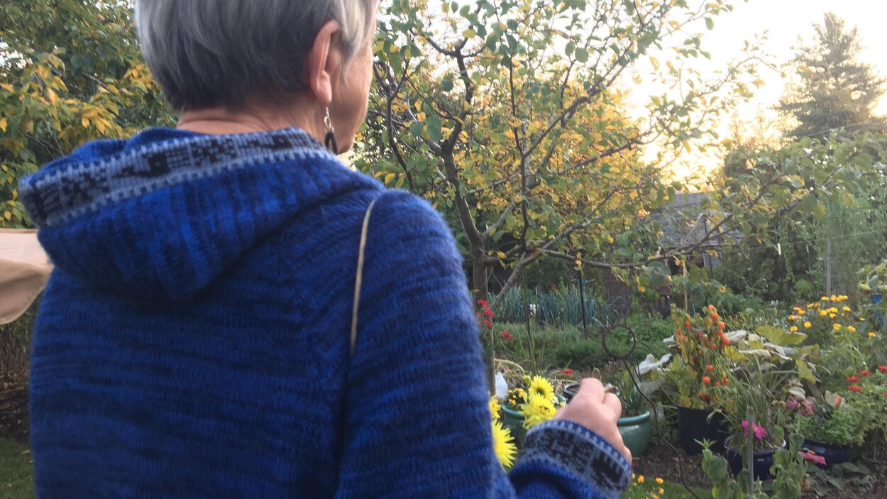 Une femme âgée tourne le dos à l'appareil photo en fumant un joint devant son jardin de fleurs.