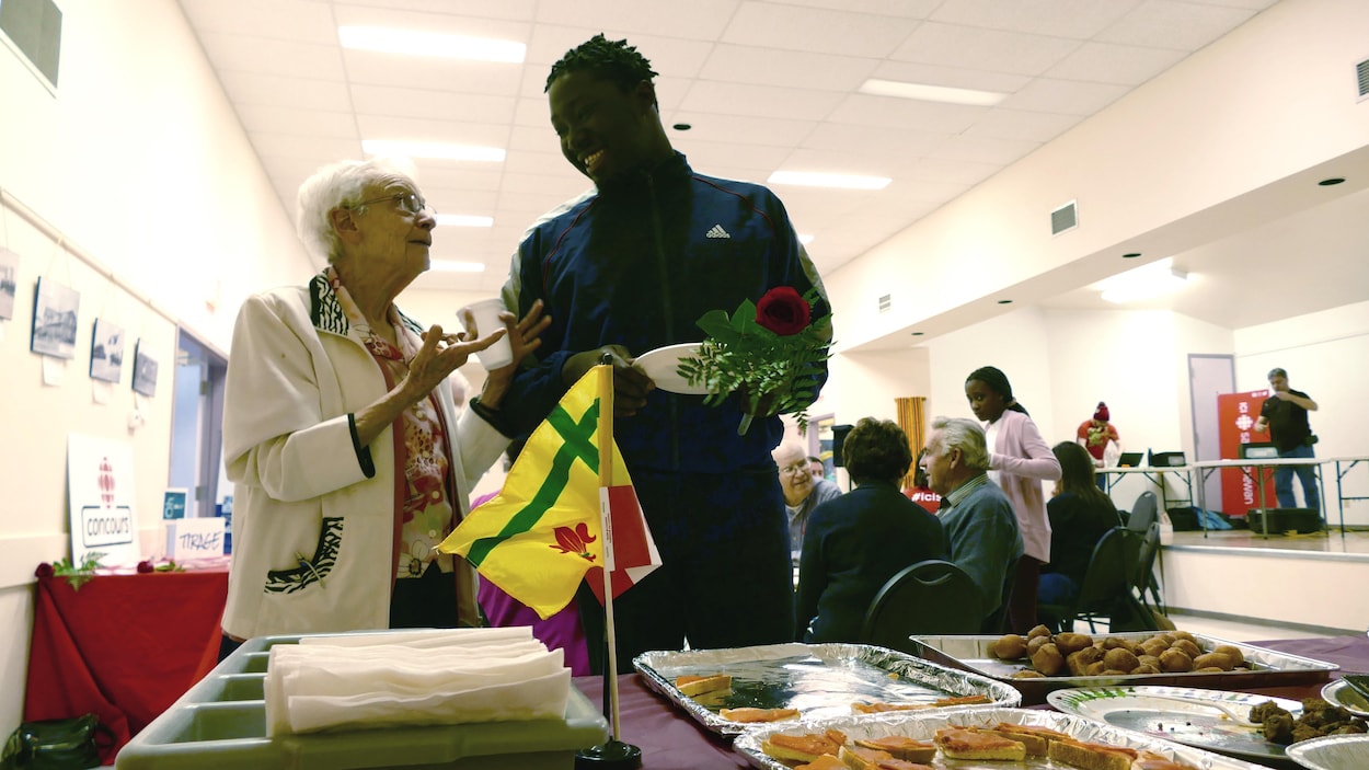 Une femme âgée et un jeune homme ont une discussion joyeuse devant un buffet. Derrière eux, des personnes sont attablées.