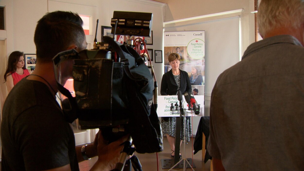 Une dame au podium devant des caméras.