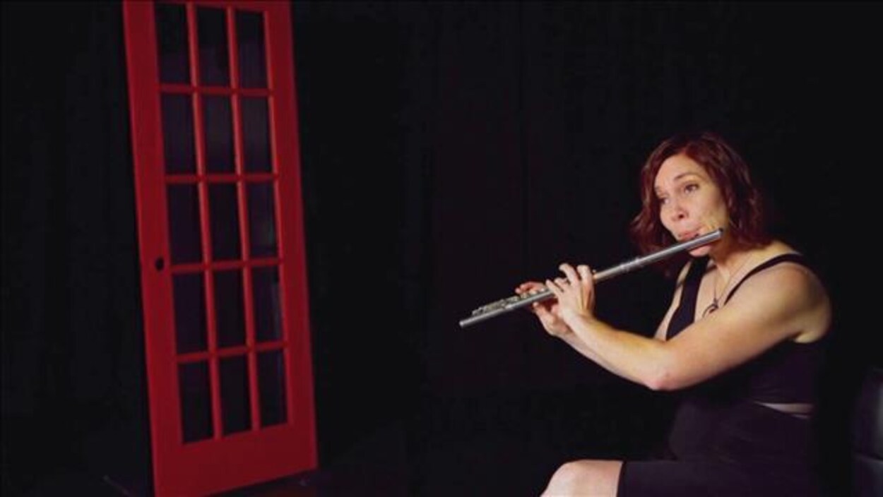 La musicienne Marie-Véronique Bourque joue de la flûte traversière dans un décor sombre contenant seulement une porte rouge.