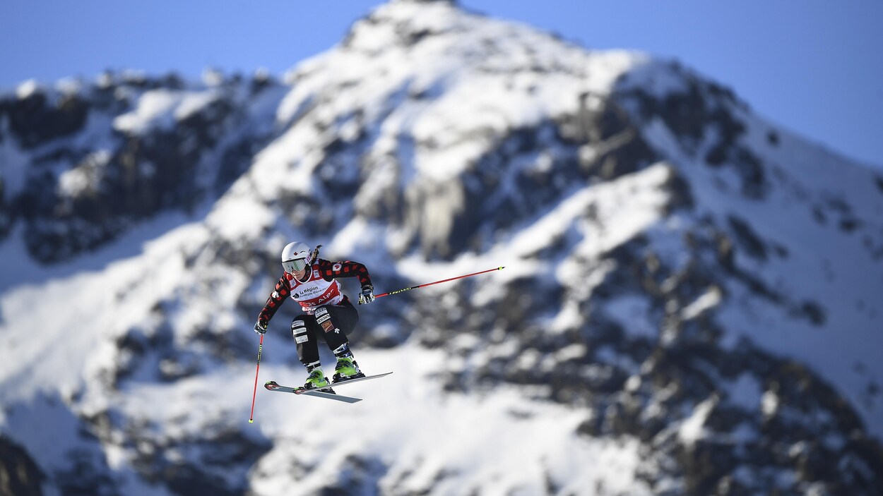 Le skieuse canadienne dans les airs pendant une épreuve de ski cross. En arrière-plan, une montagne enneigée.