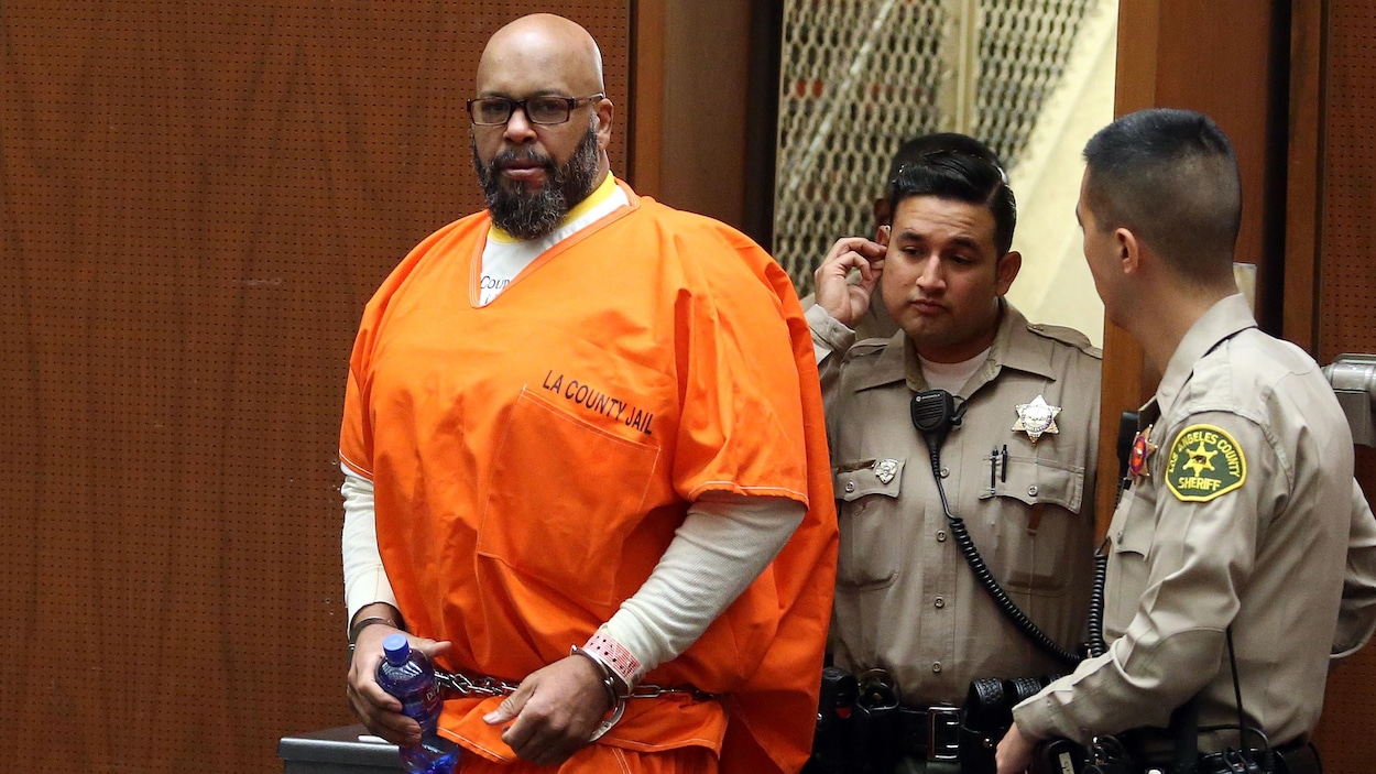 Marion «Suge» Knight, menottes aux poignets, arrive dans la salle du tribunal, regardant les objectifs des photographes. Un policier marche derrière lui. Marion «Suge» Knight, menottes aux poignets, arrive dans la salle du tribunal, regardant les objectifs des photographes. Un policier marche derrière lui.