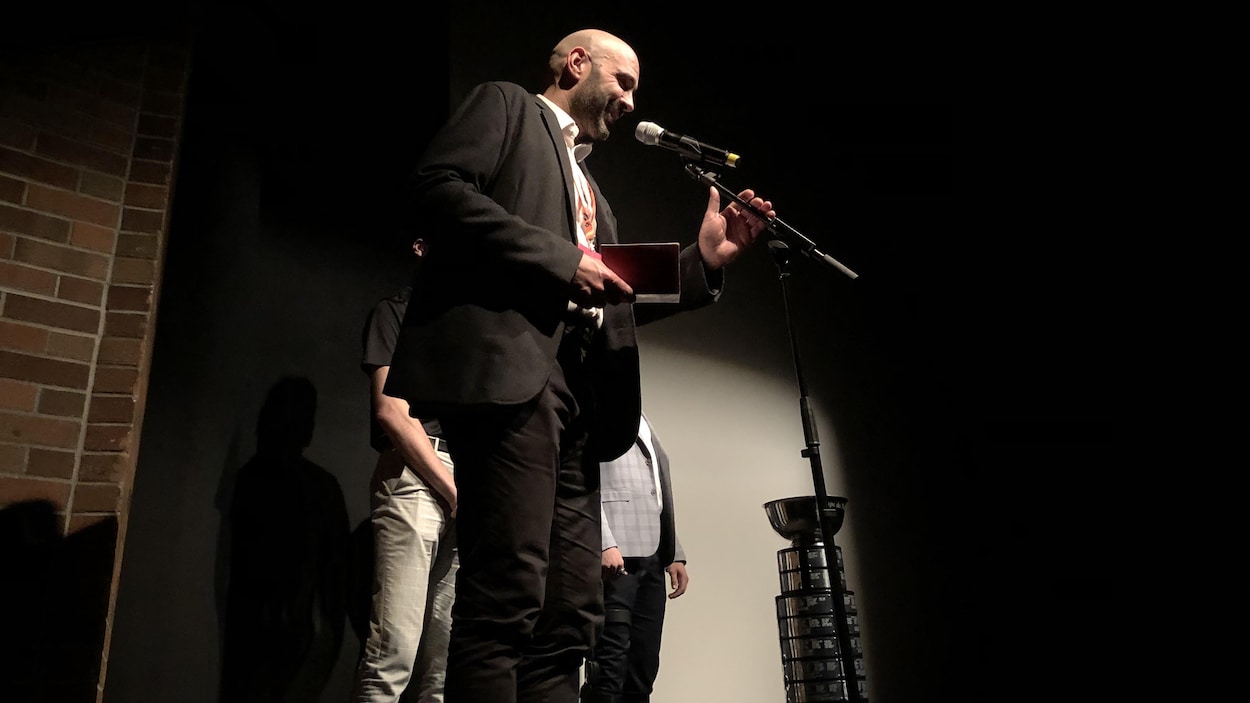 Sur une scène sombre, Martin Guérin parle au micro en tenant un petit calepin.