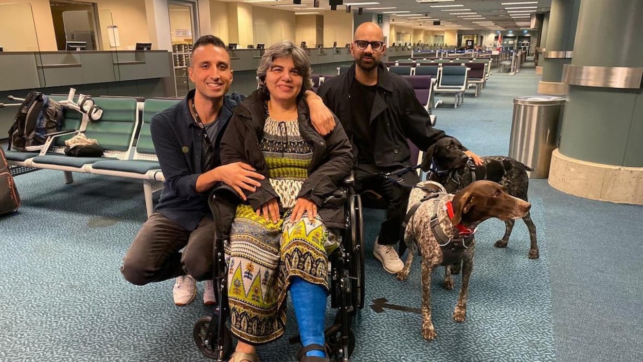 Deux hommes se tiennent aux côtés d'une femme en fauteuil roulant dans une salle d'attente d'un aéroport.