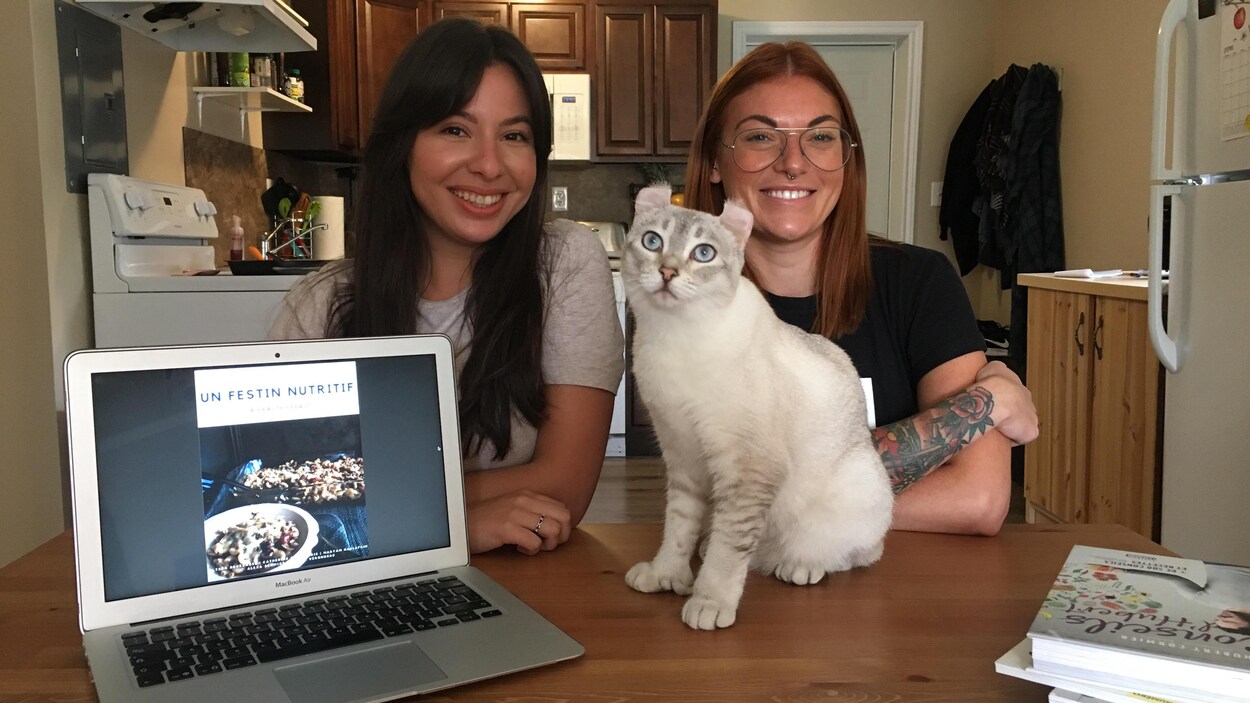 Maryam Naslafkih et Mylène Véronneau assises à une table sur laquelle se trouve un chat, un ordinateur portable et des livres de cuisine.