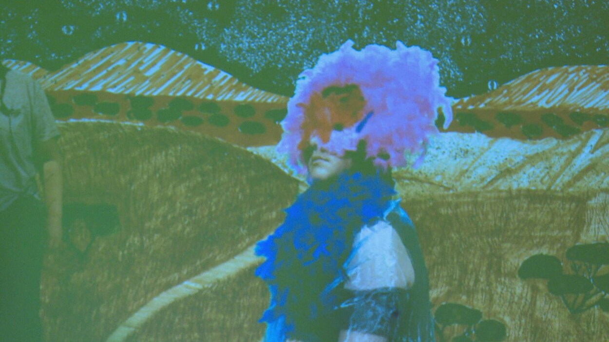 Un jeune arborant un masque à plumes roses devant un décor dessiné.