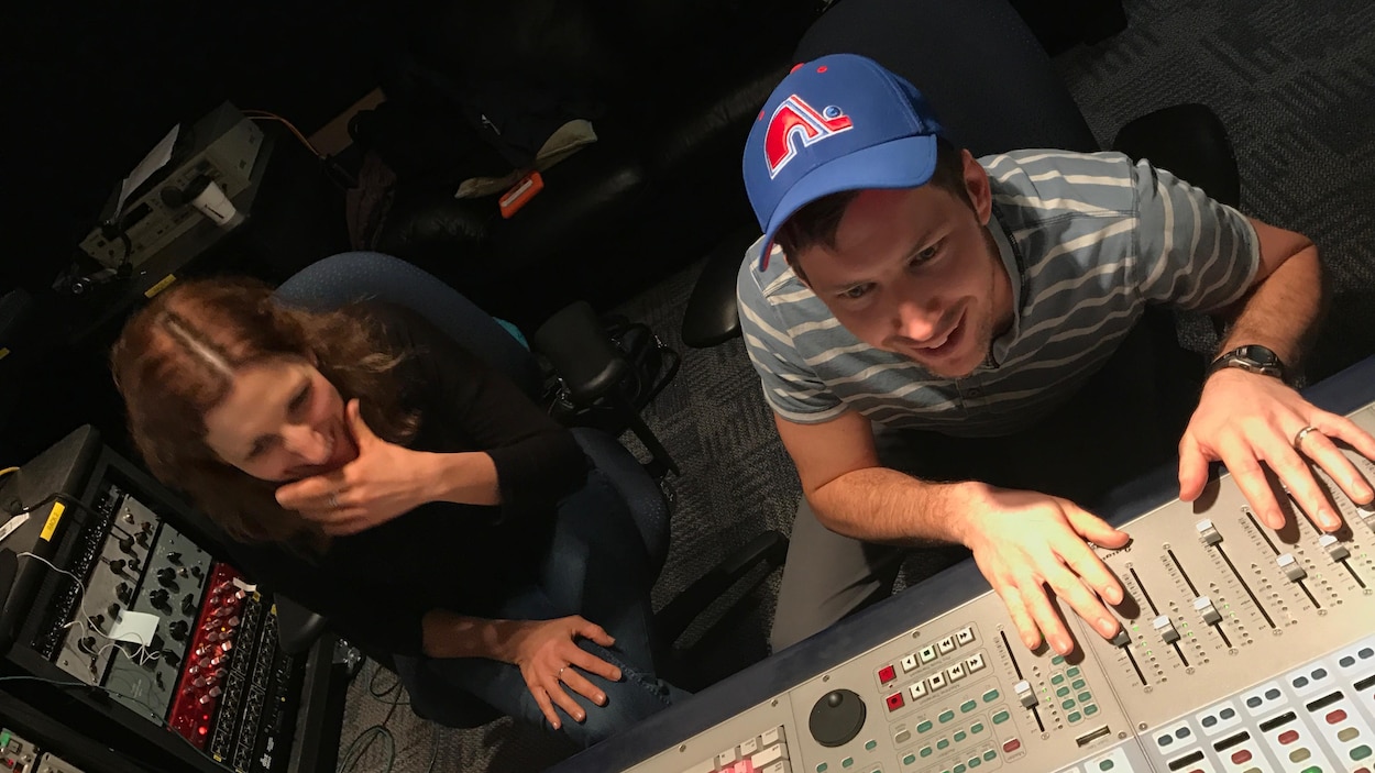 Un homme avec une casquette des Nordiques de Québec manipule des boutons d'une console en studio. À sa gauche, une femme sourit en regardant ce qu'il fait.