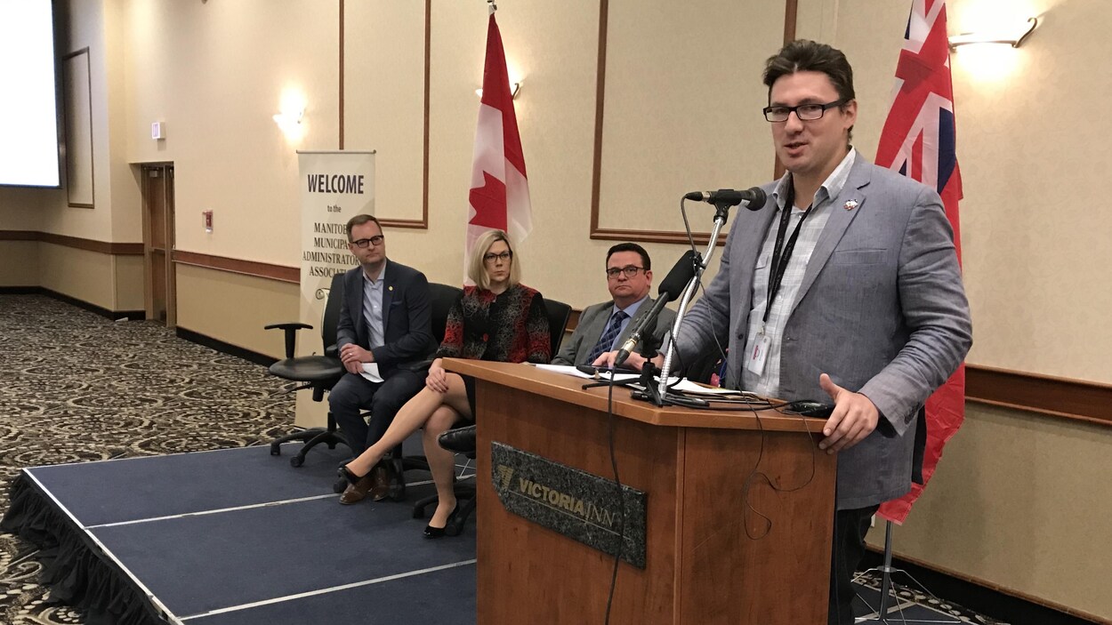 Mathieu Allard parle au micro, sur une estrade. En arrière-plan, à côté de lui, sont assis le président de l'AMM Chris Goertzen, la ministre Rochelle Squires et le ministre Jeff Wharton.