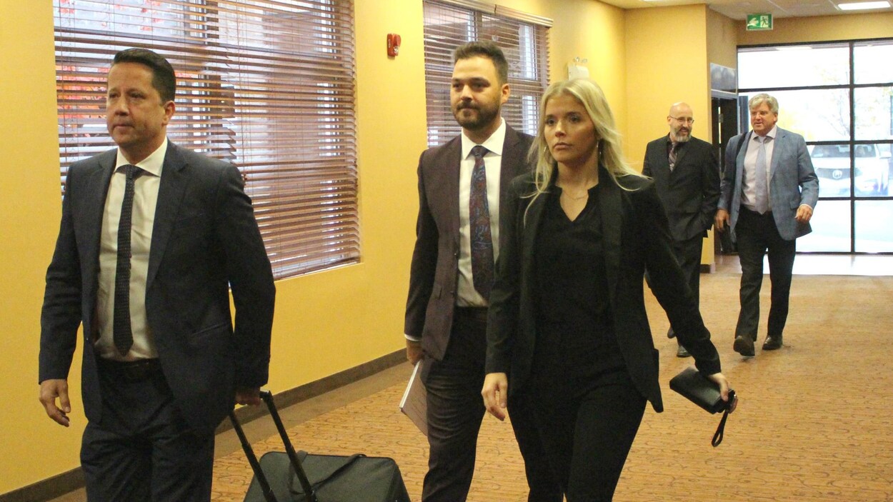 Les cinq personnes marchent dans un couloir, l'un des avocats tire une valise contenant vraisemblablement des documents.