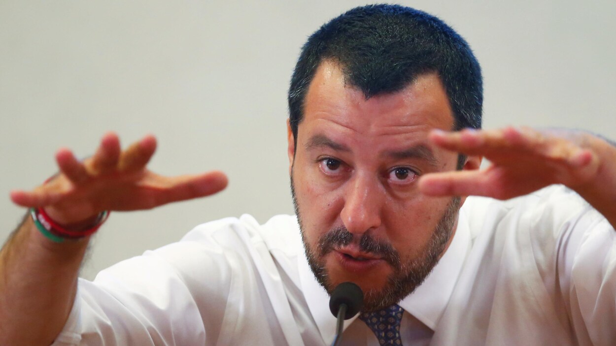 L'homme, aux cheveux courts et à la barbe fraîchement taillée, lève ses deux mains pour former une ligne virtuelle.