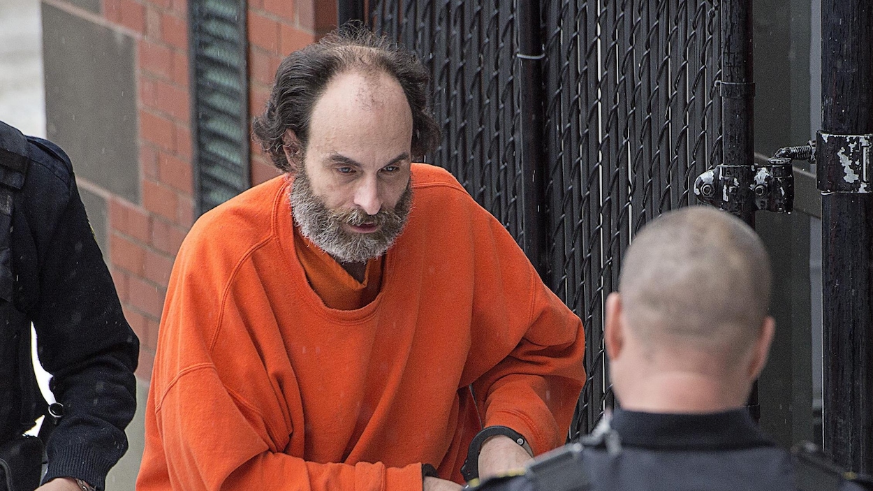 Matthew Raymond, habillé en orange, les menottes aux poignets, près d'un fourgon cellulaire.
