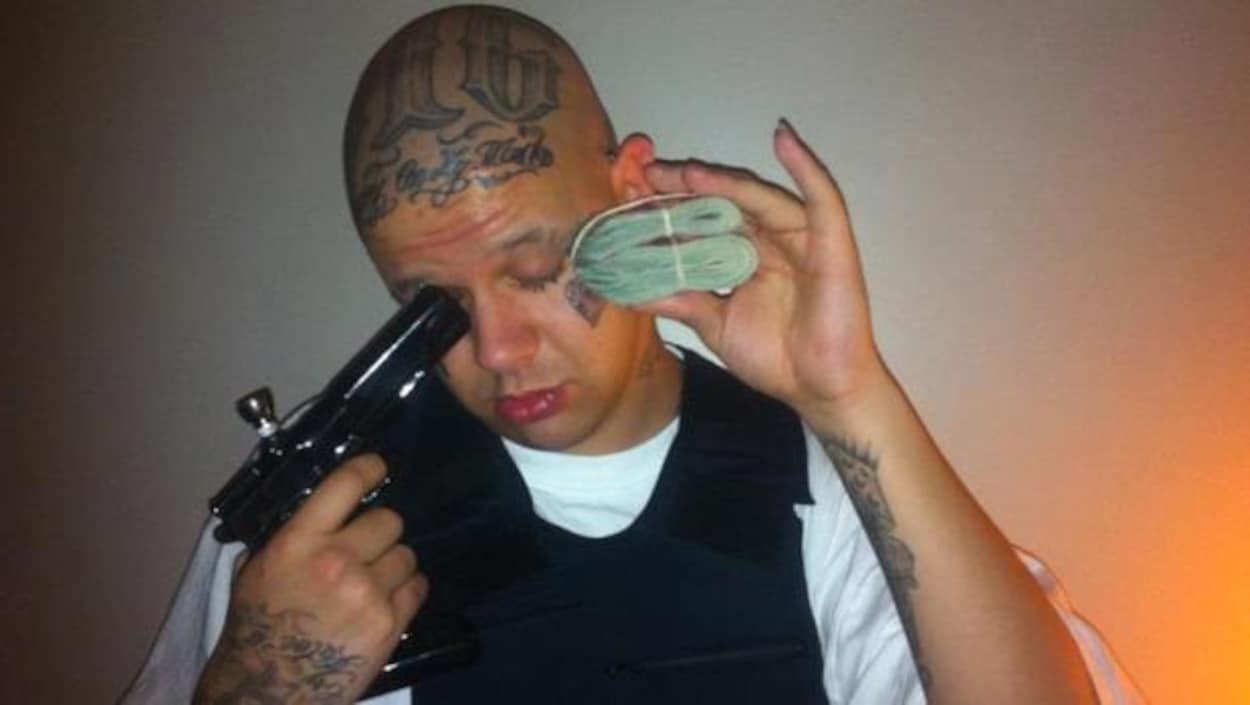 Un homme blanc avec pleins de tatouages dans le visage, pointant une fausse arme convertie en bong de marijuana vers sa figure. Il tient aussi une liasse d'argent Un homme blanc avec pleins de tatouages dans le visage, pointant une fausse arme convertie en bong de marijuana vers sa figure. Il tient aussi une liasse d'argent