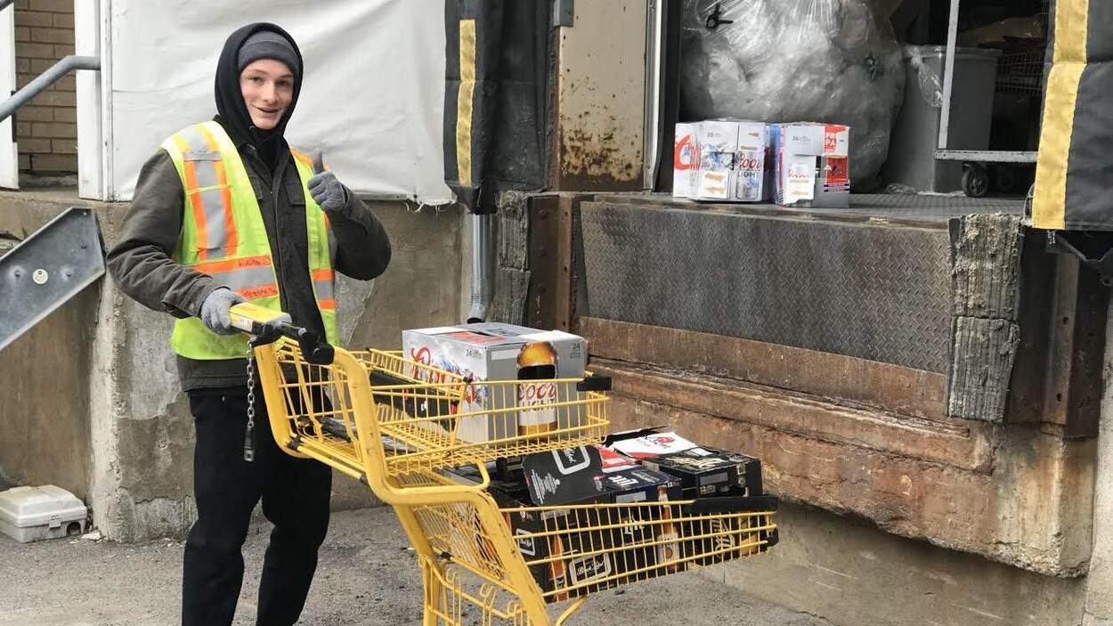 Un jeune homme tient un panier d'épicerie jaune avec des caisses de bières dedans. Il sourit et tient son pouce dans les airs.