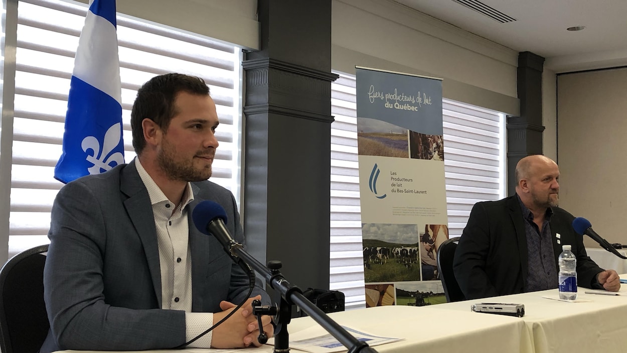 Les deux hommes sont assis à une table de conférence. Ils ont tous les deux un micro devant eux et regardent le public.