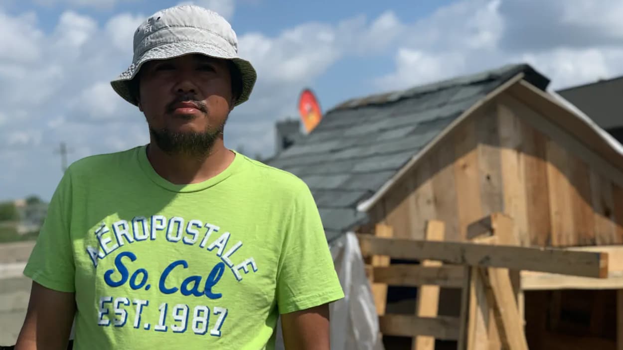 Un homme devant une cabane de fortune faite de bois avec une toiture en bardeaux d'asphalte.