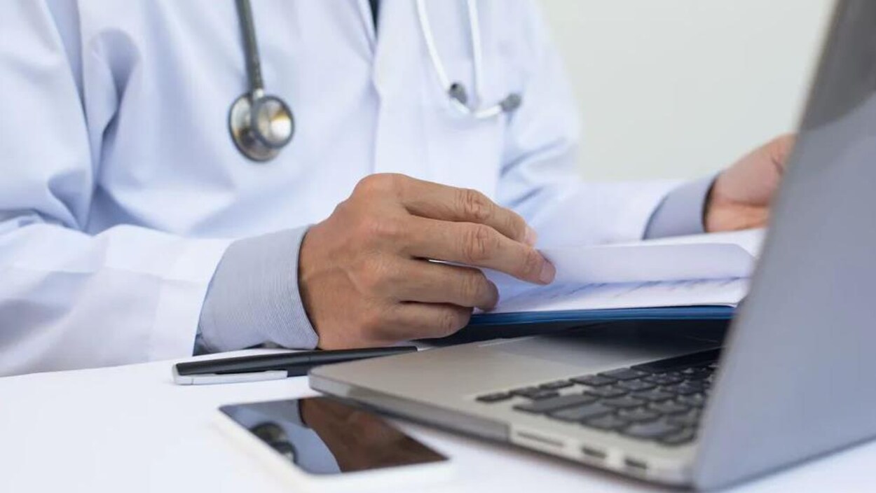 Un médecin avec un stéthoscope autour du cou regarde ses notes dans un cahier en face de son ordinateur portable et de son cellulaire.