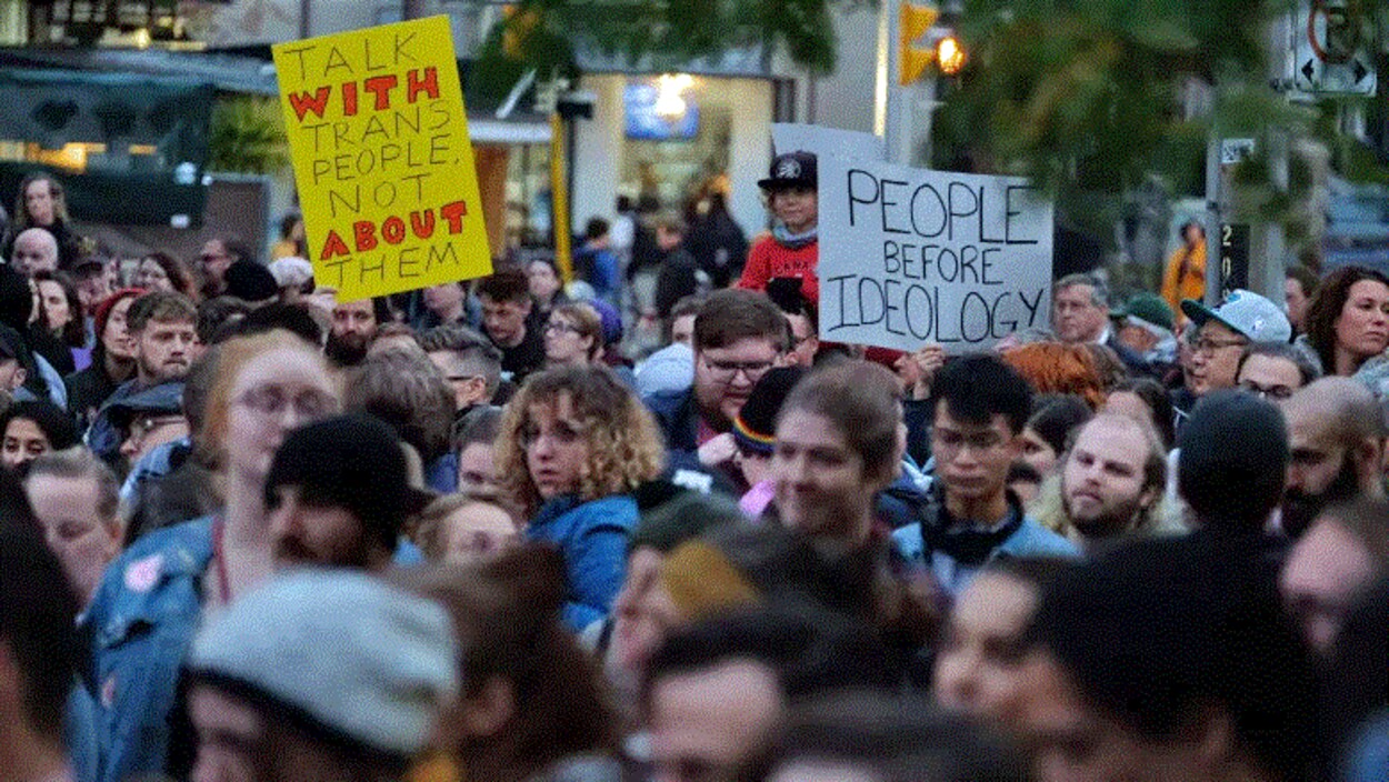 Des manifestants tiennent des affiches avec des messages comme « parle avec les personnes trans, pas à leur sujet ».