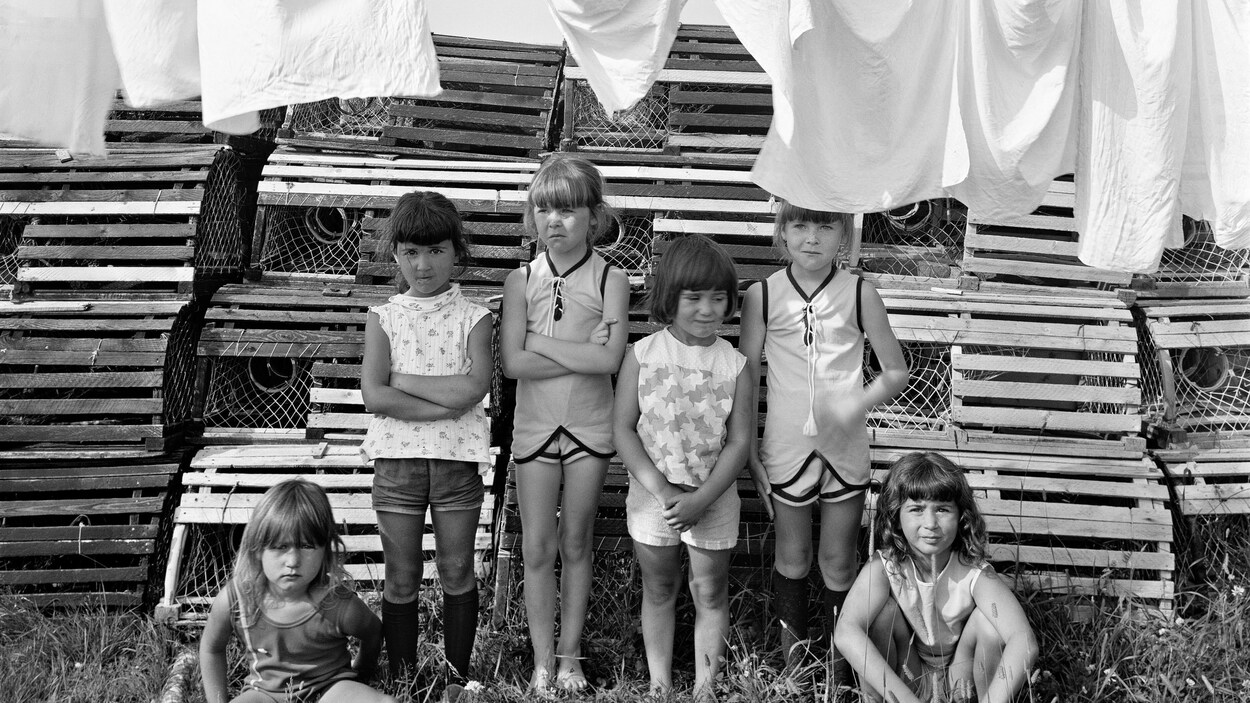 Quatre fillettes debout et deux assises dans l'herbe, entre une pile de casiers à homard et des vêtements blancs sur une corde à linge.