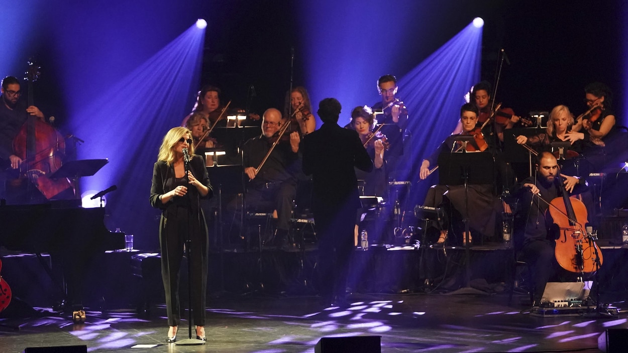 La chanteuse, qui porte des verres fumés, chante au micro. Derrière elle, un ensemble de cordes l'accompagne.