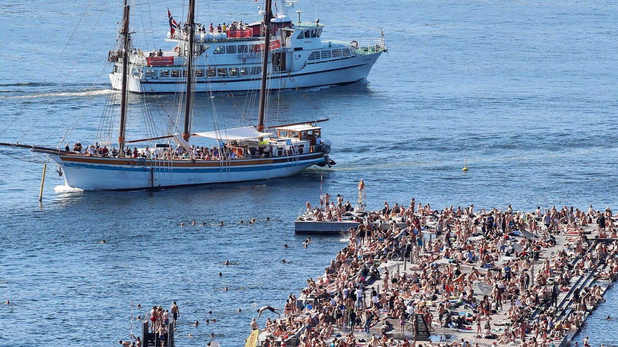 Une plage publique bondée de monde en Norvège où un grand voilier et un bateau de croisière sont visibles.