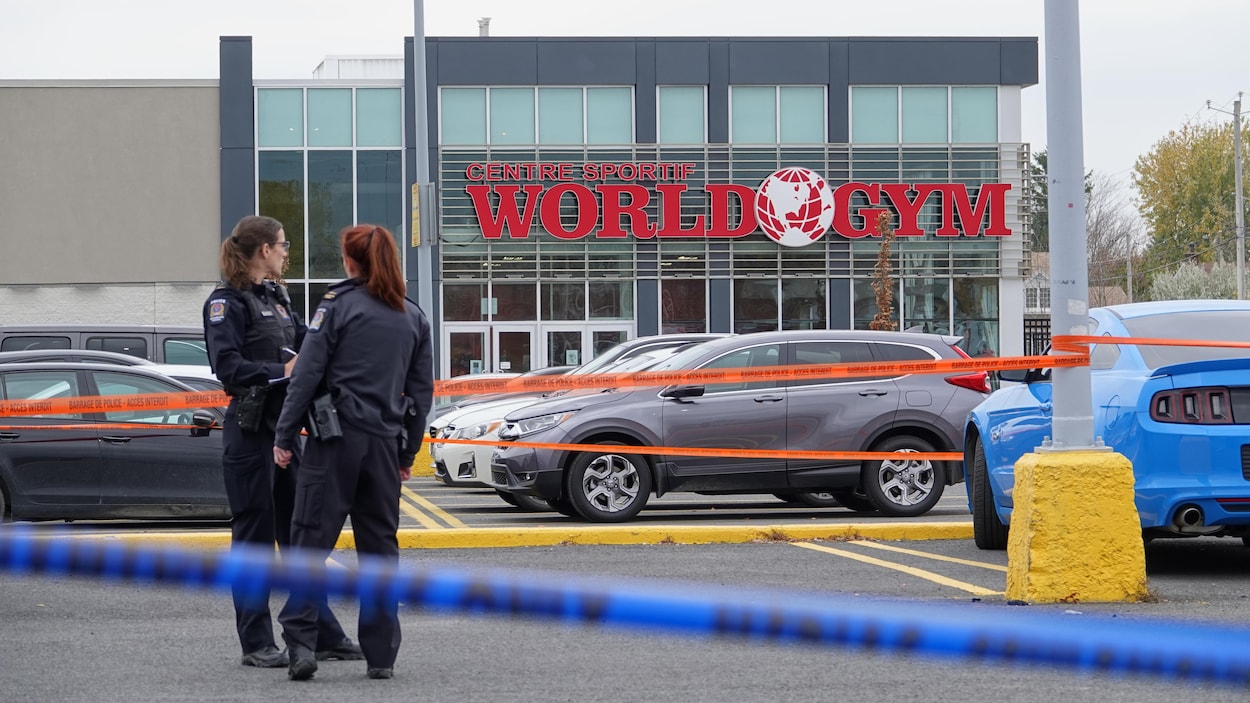Deux policières discutent derrière un cordon de sécurité, dans un stationnement, devant la devanture du centre sportif World Gym.