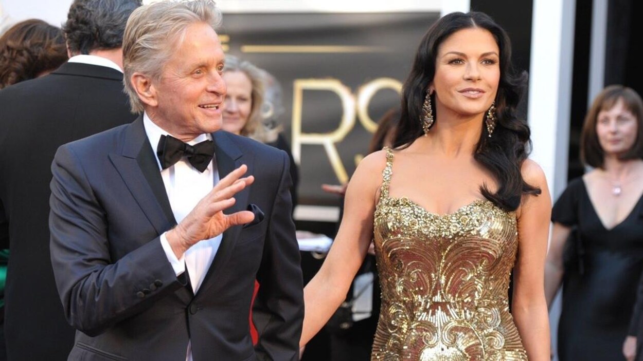 Michael Douglas et Catherine Zeta-Jones, habillés en tenue de soirée (costume bleu pour lui et robe dorée pour elle) se tiennent par la main sur un tapis rouge en faisant signe aux public. Michael Douglas et Catherine Zeta-Jones, habillés en tenue de soirée (costume bleu pour lui et robe dorée pour elle) se tiennent par la main sur un tapis rouge en faisant signe aux public.