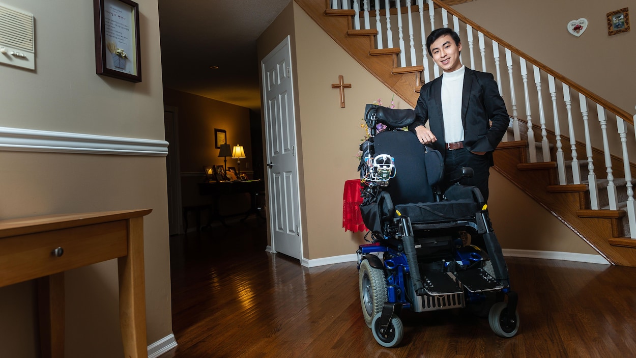 Michael Ko se tient debout à côté d'un fauteuil roulant électrique dans un hall d'entrée résidentiel.