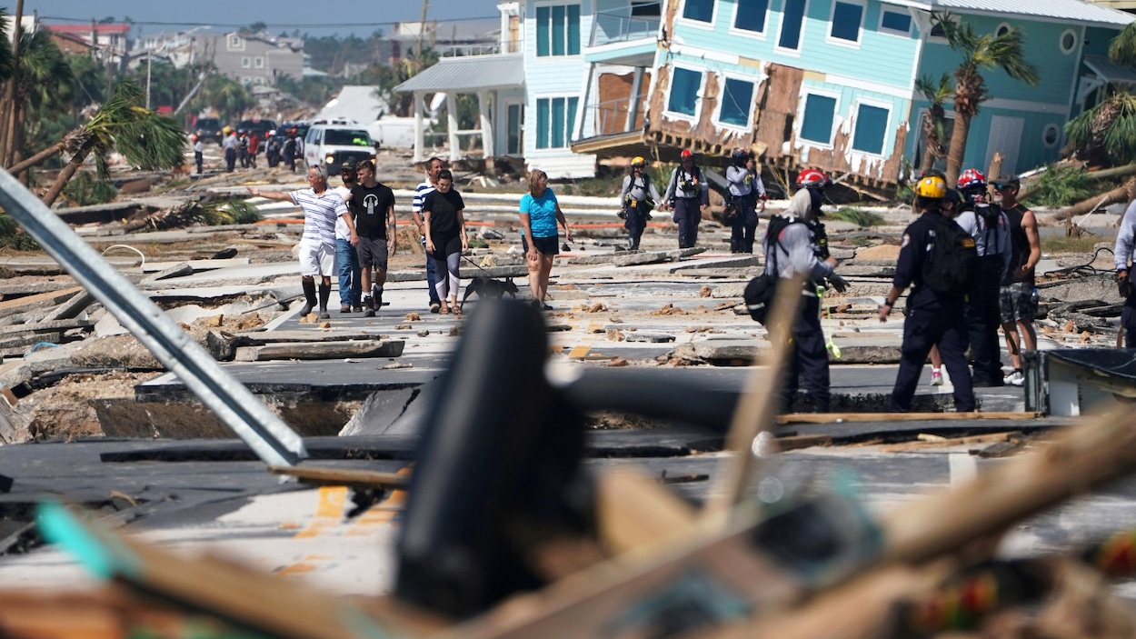 Secouristes et premiers répondants marchent dans une rue jonchée de débris. Des maisons sont lourdement endommagées. 