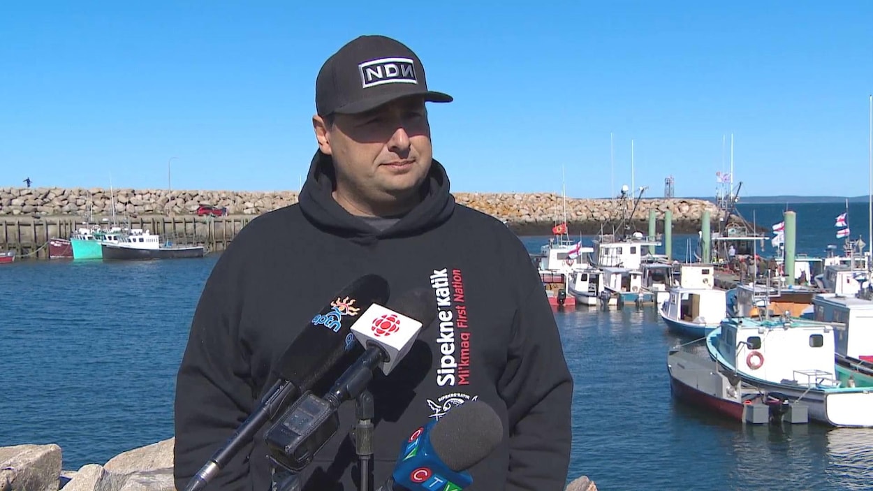 Mike Sack sur le quai, dos à une flotte de bateaux, devant plusieurs micros de différents médias.