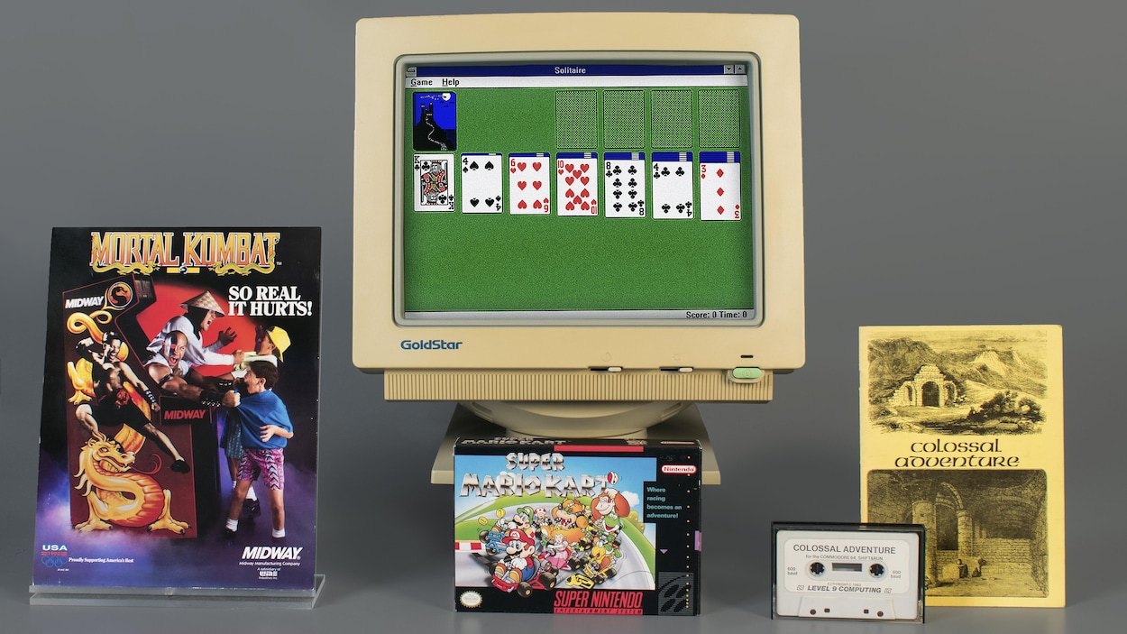 Un photo montrant une affiche pour la borne d'arcade de Mortal Kombat, la pochette américaine du jeu Super Mario Kart, une cassette audio et un livret jaune de Colossal Cave Adventure et un vieil écran d'ordinateur affichant une vieille version du jeu Microsoft Solitaire.