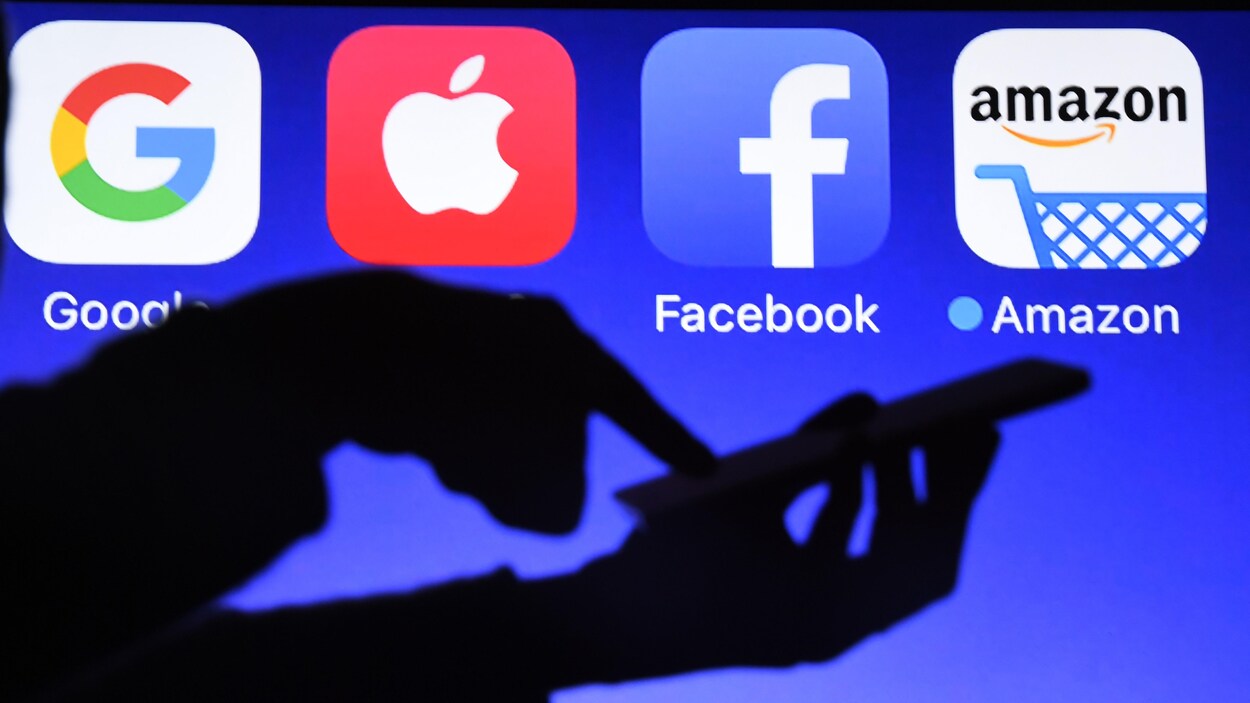 Une personne utilise un téléphone cellulaire devant un écran affichant des logos de Google, Apple, Facebook et Amazon.