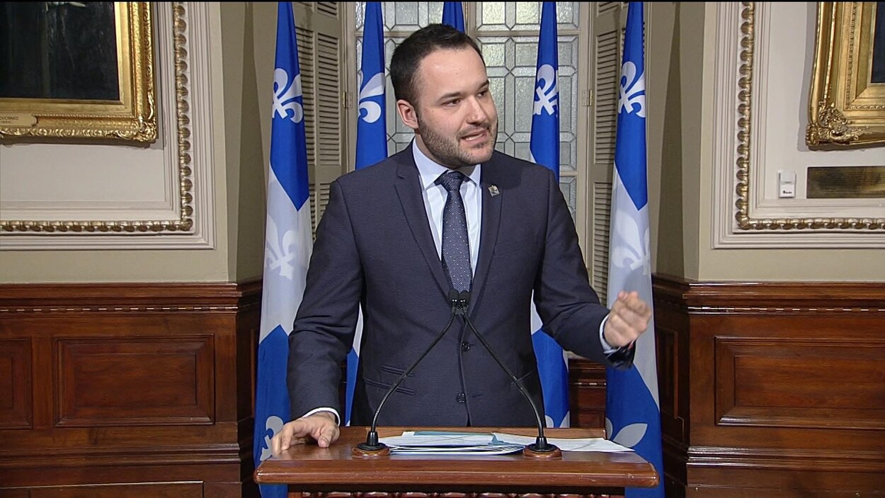 Mathieu Lacombe s'adresse aux journalistes à un podium, devant des drapeaux du Québec.