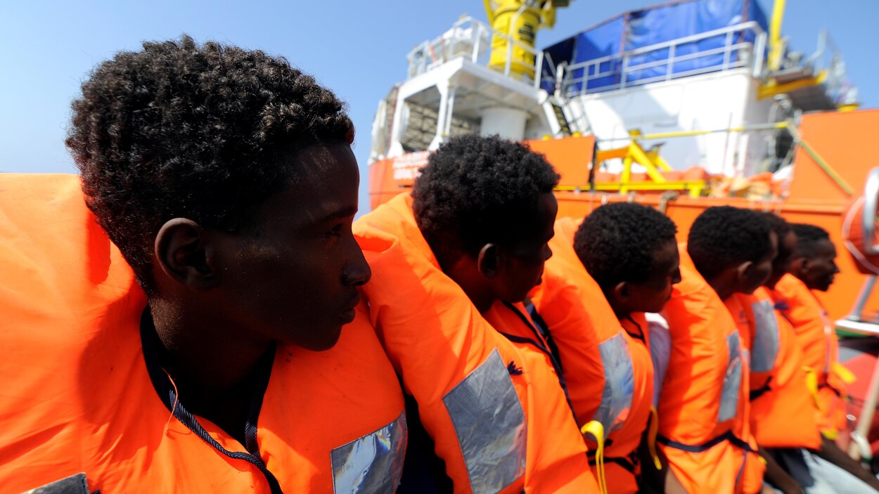 Des migrants vêtus de gilets de sauvetage attendent de monter sur l'Aquarius qui se trouve derrière eux. Des migrants vêtus de gilets de sauvetage attendent de monter sur l'Aquarius qui se trouve derrière eux.