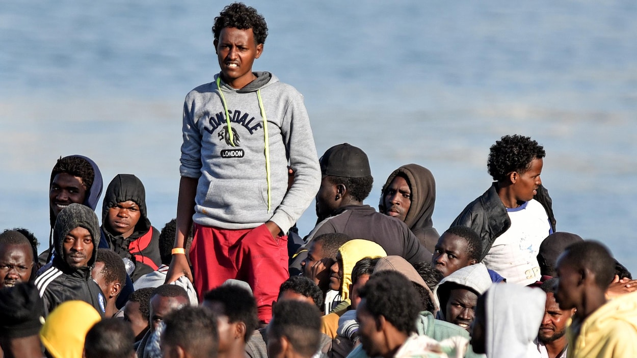 Plusieurs hommes attendent de descendre d'un bateau. L'un d'eux surplombe les autres.