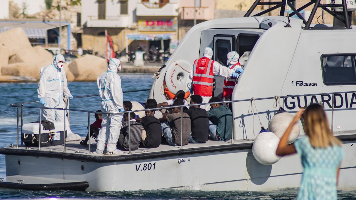 De jeunes hommes sont assis sur le pont d'un petit bateau, entourés de gens vêtus de masques et de combinaisons qui les couvrent des pieds à la tête.