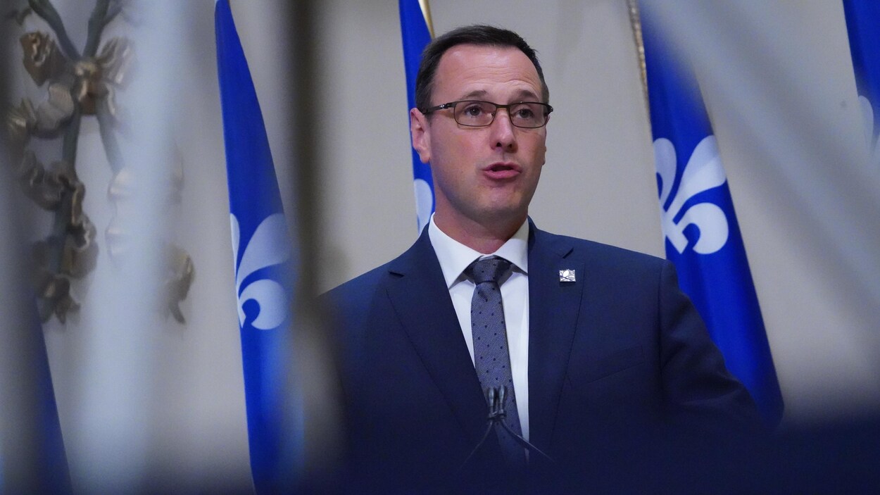 Il donne un point de presse avec des drapeaux du Québec derrière lui.