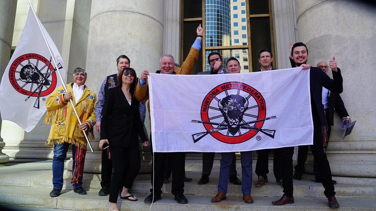 Un groupe de personnes qui sourient en tenant des drapeaux devant un bâtiment avec des colonnes.