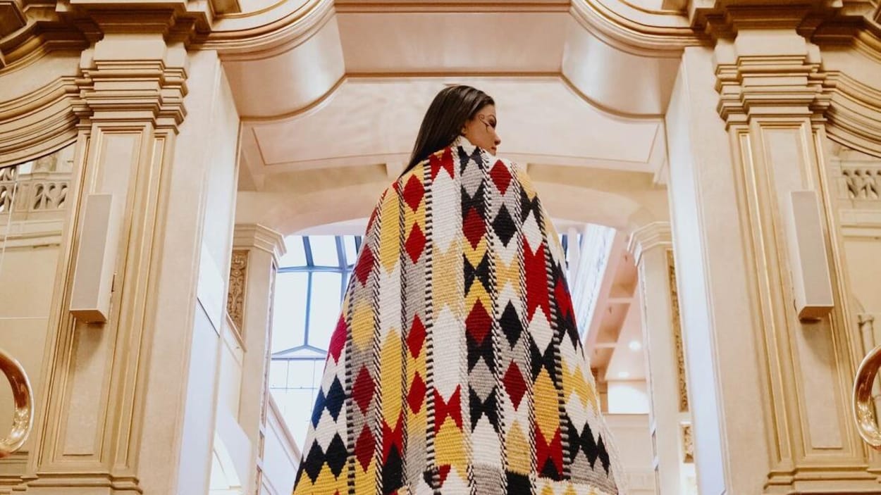 Une femme, de dos, porte une cape multicolore aux motifs autochtones.